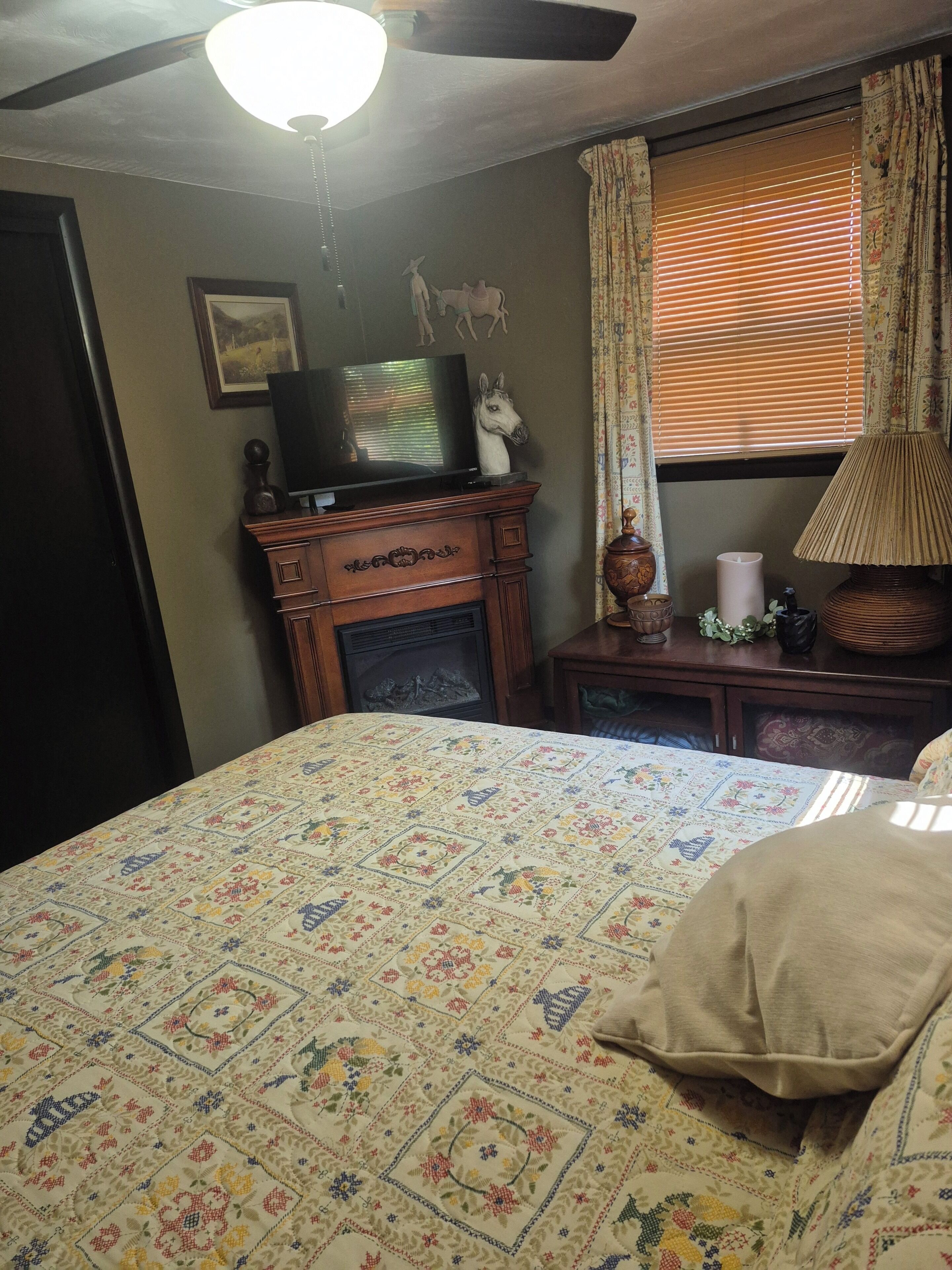 Un dormitorio con cama, televisión sobre un mueble de madera, chimenea, lámpara y ventana con cortinas.