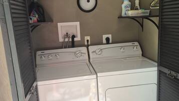 A laundry room with two stacked washers and dryers, a wall-mounted light fixture, and a shelf with cleaning supplies.