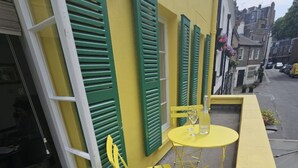 A yellow outdoor seating area with a table and chairs, a window with green shutters, and a yellow awning.