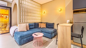 Un salon moderne avec un canapé d’angle, une petite table ronde, une table d’appoint en bois et un coin cuisine intégré.