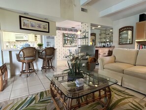 A spacious living room with a glass coffee table, a beige sofa, and a dining area with wooden chairs and a kitchen in the background. - Cozy home, pet friendly, private pool & close to beach. Minimum 60 days (Osprey)