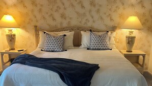 A neatly made bed with a white duvet and black throw, flanked by patterned pillows, in a room with floral wallpaper and two bedside lamps.