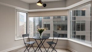 A corner room with a round table and two chairs, a vase with flowers, and large windows with city views.