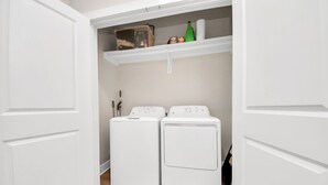 Une buanderie avec deux machines à laver à chargement frontal, de couleur blanche, et une étagère au-dessus, contenant une bouteille verte, une boîte et divers autres objets.