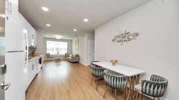 Une cuisine moderne avec des meubles blancs, un coin repas avec une table blanche et des chaises à revêtement gris, et un salon avec un canapé et des fauteuils.