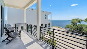 A balcony with two Adirondack chairs, a view of the sea, and a white building in the background. - Stylish Oceanview Kayak Getaway w/ Pool & Hot Tub! (Key Largo)