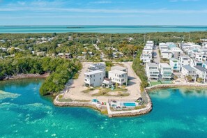 An aerial view of a coastal resort with modern buildings, a central pool area, and clear turquoise waters. - Stylish Oceanview Kayak Getaway w/ Pool & Hot Tub! (Key Largo)