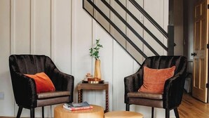 Una habitación con dos sillas de mimbre negro, una pequeña mesa de madera, una escalera y una puerta que da a otra habitación.