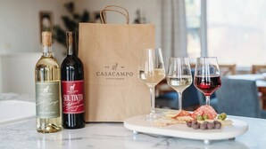 Una mesa con dos botellas de vino, una bolsa de papel, tres copas de vino y una tabla de quesos.