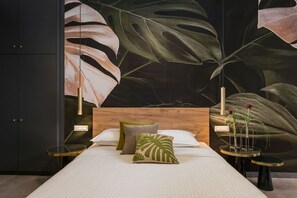 Un dormitorio con cabecera de madera, cama con ropa blanca, almohadas verdes y beige, una mesita de vidrio con una vela y un mural de hojas grandes en la pared.