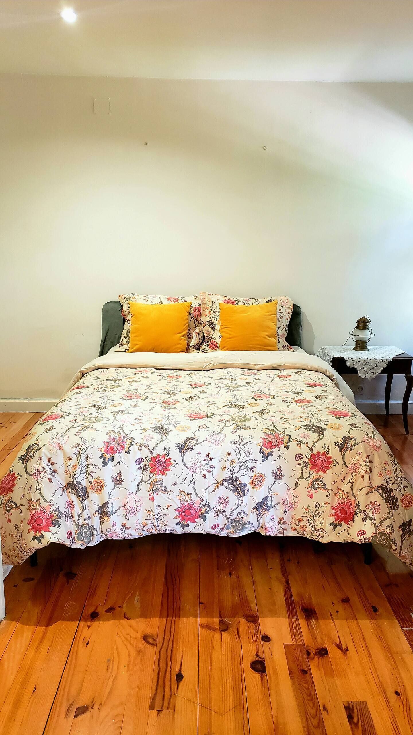 Un dormitorio con colcha estampada, dos almohadas amarillas, suelo de madera, una mesita de noche con lámpara y una silla.