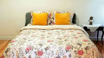 Un dormitorio con colcha estampada, dos almohadas amarillas, suelo de madera, una mesita de noche con lámpara y una silla.