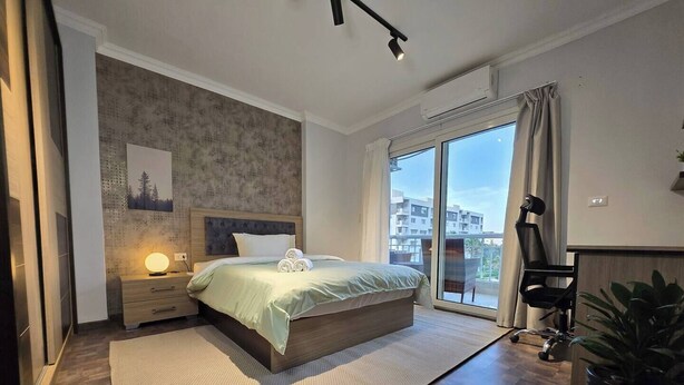 A modern bedroom with a large bed, a nightstand, a wall-mounted lamp, a sliding glass door to a balcony, and a potted plant. - Madinaty Botanic Bliss (Cairo Governorate)