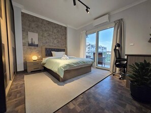 A modern bedroom with a large bed, a nightstand, a wall-mounted lamp, a sliding glass door to a balcony, and a potted plant. - Madinaty Botanic Bliss (Cairo Governorate)