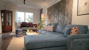 A modern living room with a sectional sofa, a dining area with a wooden cabinet, and a large window providing natural light. - Madinaty Botanic Bliss (Cairo Governorate)