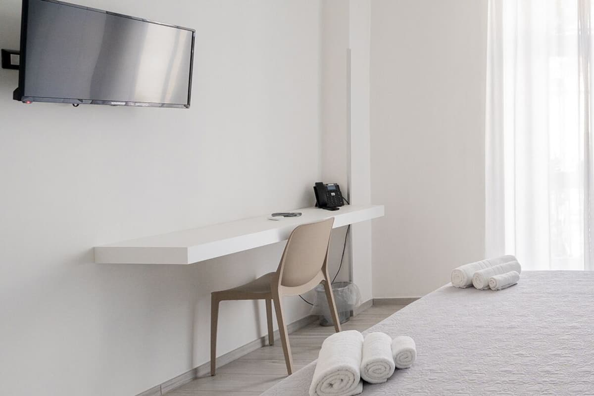 A hotel room with a bed, a desk with a chair, a TV mounted on the wall, and a window with sheer curtains.