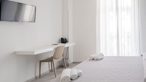 A hotel room with a bed, a desk with a chair, a TV mounted on the wall, and a window with sheer curtains. - Harmonia home (Capitello)