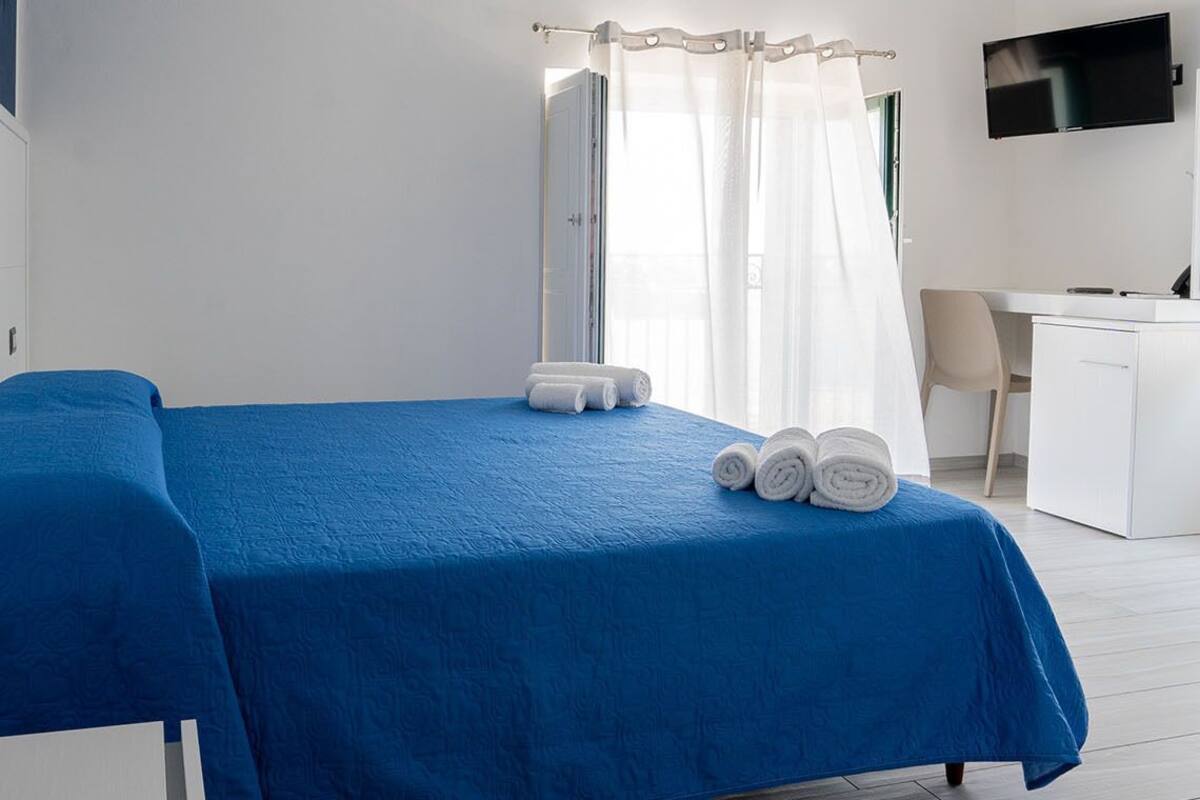 A room with a bed covered in blue bedding, a small desk with a chair, a TV mounted on the wall, and a shower area with curtains.