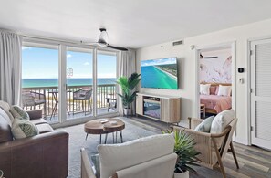 A spacious living room with a large TV, comfortable seating, and a balcony with a view of the ocean.