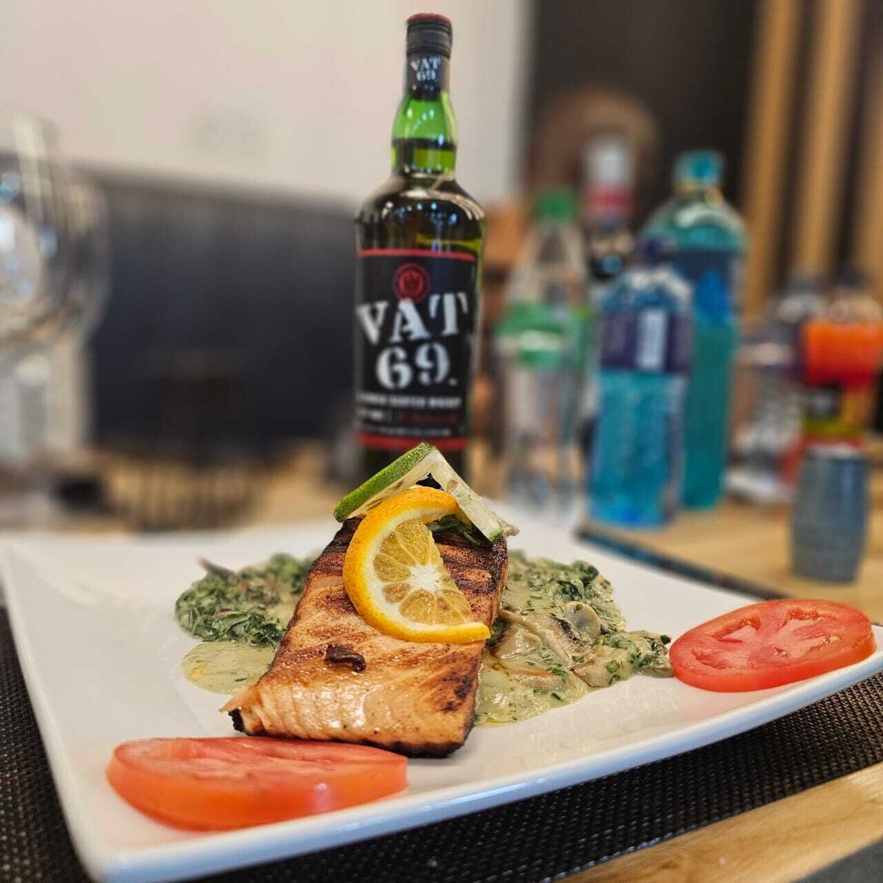 A plate of grilled salmon with lemon and herb garnish, accompanied by a bottle of V&T 69 whiskey, and a glass of water on a table.