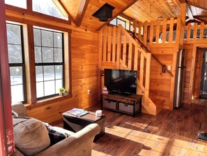 L’intérieur d’un chalet chaleureux, avec des murs en bois, un téléviseur posé sur un meuble en bois, un canapé et un escalier.