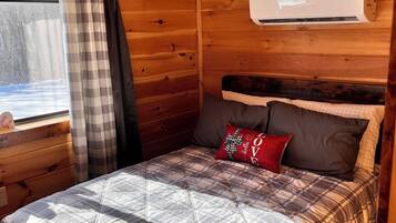 Ein gemütliches Zimmer in einer Holzhütte mit einem Bett, einem Fenster mit Vorhängen und einer Klimaanlage.