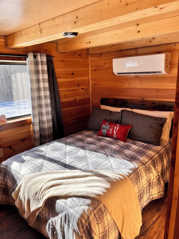 A cozy wooden cabin room with a bed, a window with curtains, and an air conditioning unit.