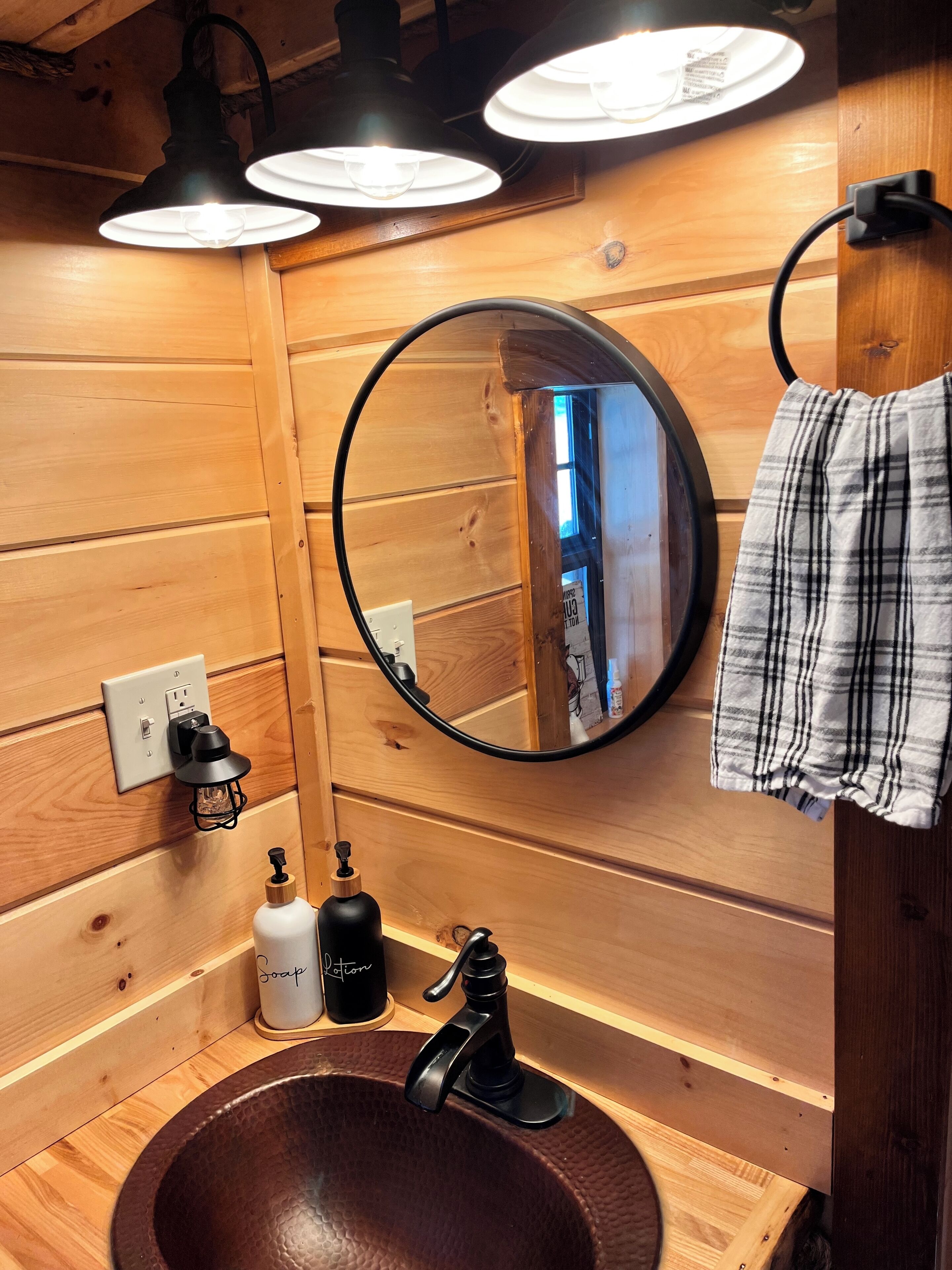 A wooden-paneled bathroom with a round mirror, a round sink, and three pendant lights.