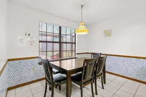 Ein Essbereich mit Holztisch und Stühlen, einer Hängelampe, einem Fenster mit Jalousien und einem Fliesenboden.