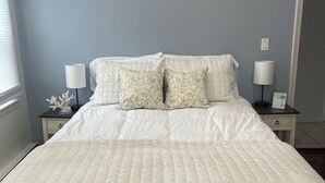 Cama bem arrumada com um edredom branco acolchoado e duas almofadas estampadas, acompanhada por duas mesas de cabeceira com luminárias e um pequeno objeto decorativo.