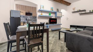 A modern living room with a dining area, a kitchen with wooden cabinets, and a TV displaying "NETFLIX". - ROSA HOMES - THE GREEN  (Genoa)