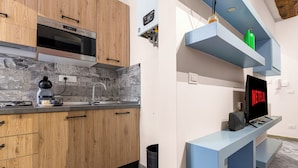 A modern kitchen with wooden cabinets, a built-in microwave, and a sink. There is a black trash can and a blue entertainment unit with a TV displaying "NETFLIX". - ROSA HOMES - THE GREEN  (Genoa)