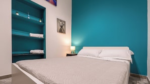 A bedroom with a bed, teal shelving unit, and a nightstand with a lamp. - ROSA HOMES - THE GREEN  (Genoa)