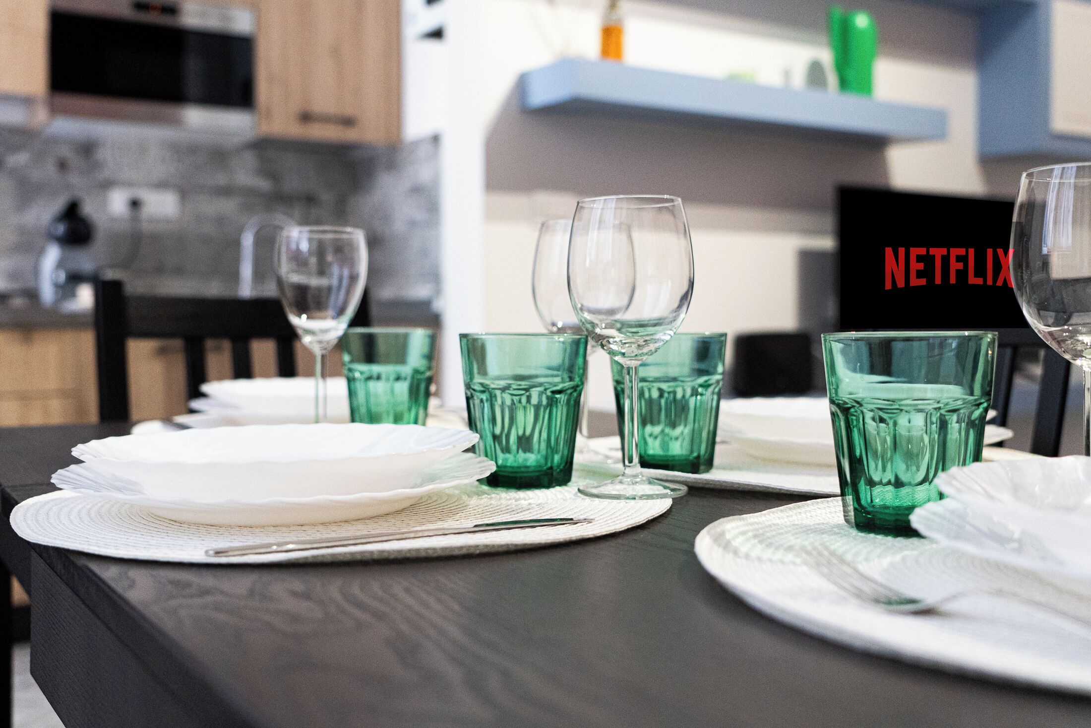 Una mesa de comedor dispuesta con vasos verdes y platos blancos, con un letrero de Netflix al fondo.
