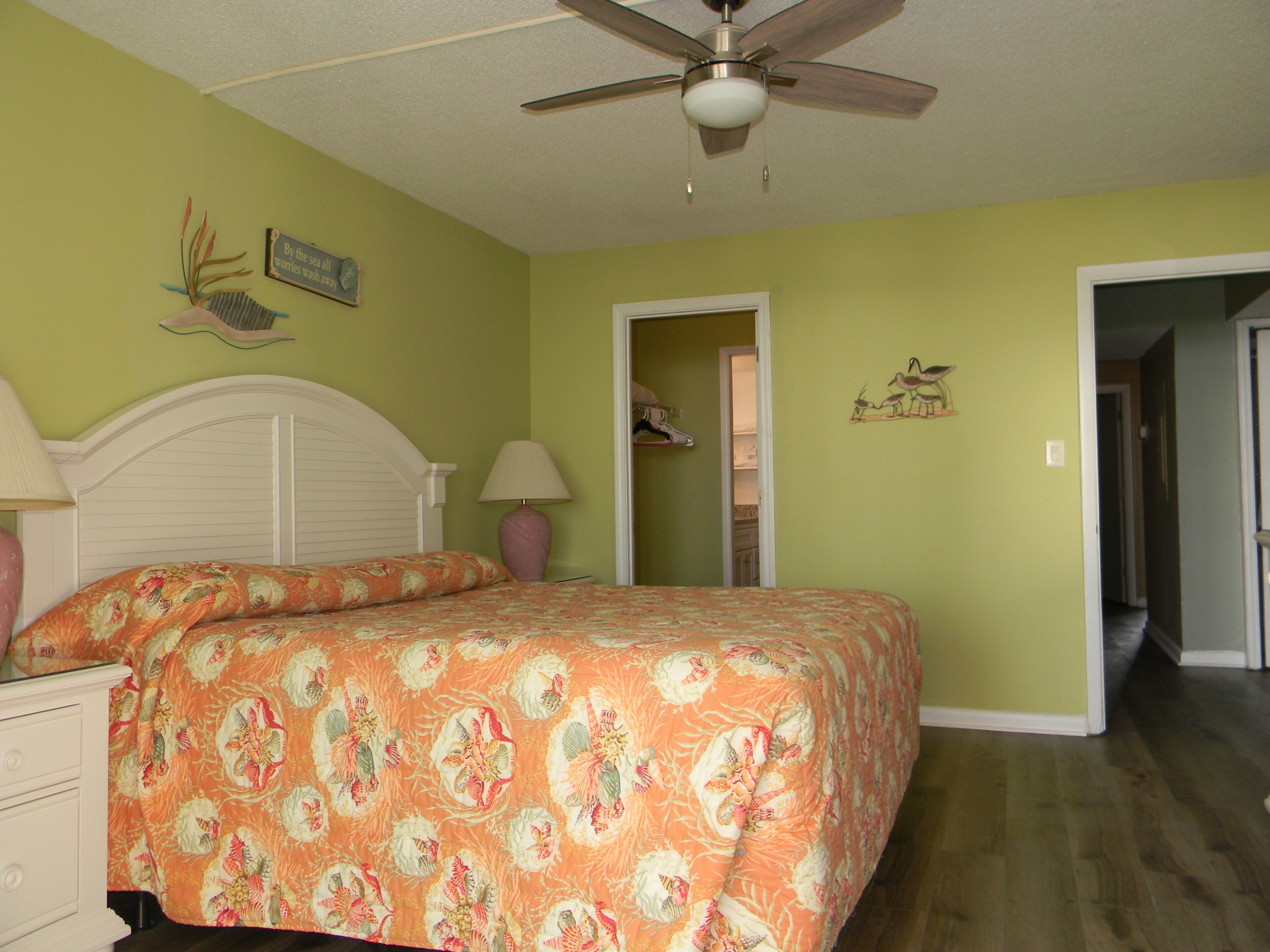 A bedroom with a bed, a nightstand, a ceiling fan, and a door leading to another room.