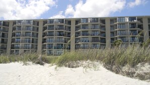 A multi-story hotel with balconies and large windows, situated on a sandy beach with overcast sky.
