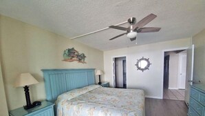 A bedroom with a bed, a nightstand, a ceiling fan, a wall clock, and a door leading to another room. - Oceanfront Three Bedroom Condo - Crescent Sands C-4-September discounted! (North Myrtle Beach)