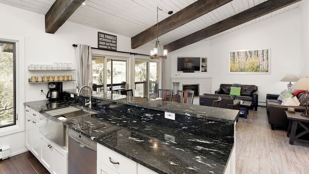A modern kitchen with a large island, white cabinetry, and a wooden ceiling beam. There is a dining area with a fireplace and a sofa. - Beautiful 2 Bedroom + Loft Located in Snowmass (Snowmass Village)