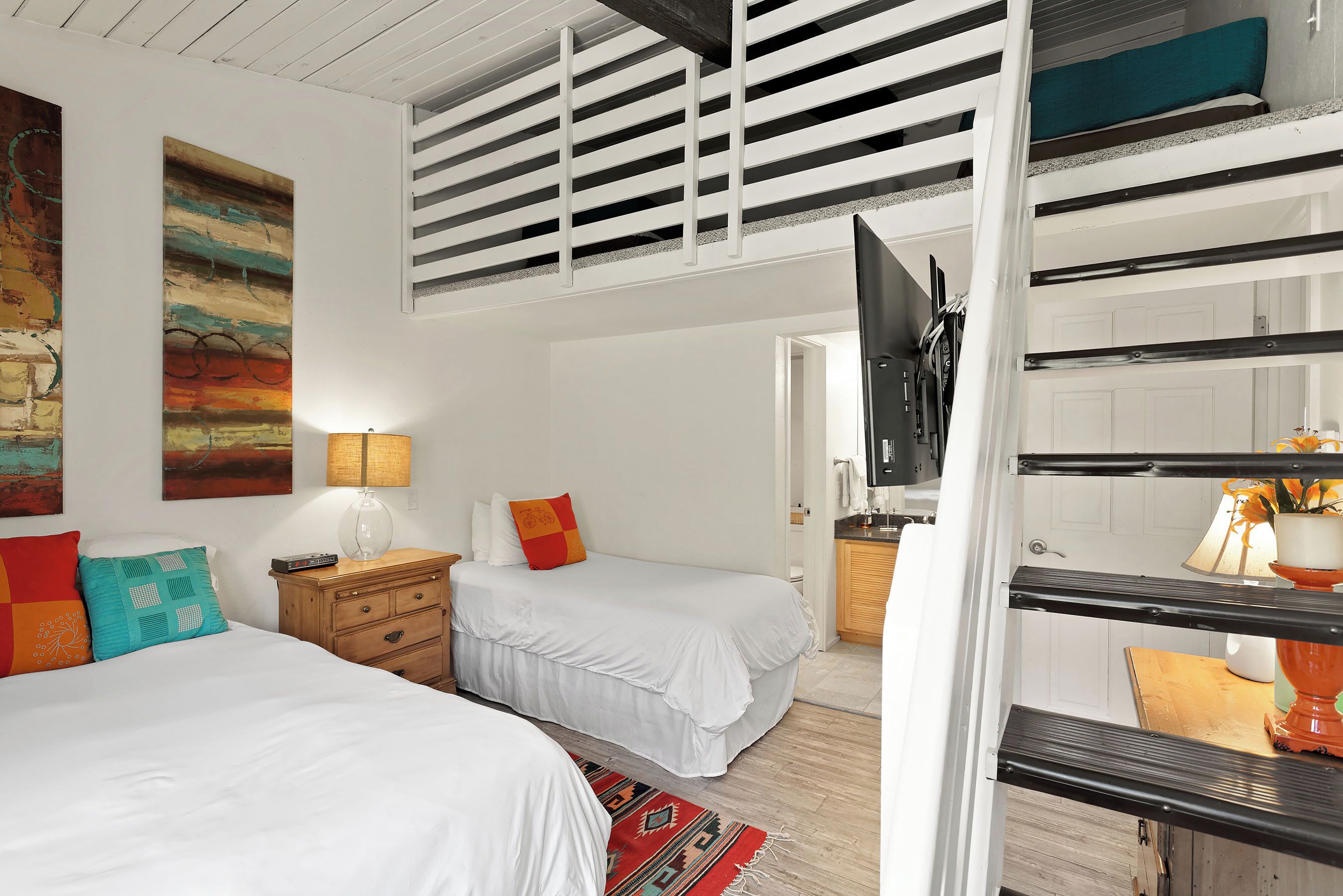 A bedroom with two beds, a wooden dresser, a TV, and a kitchen area visible through an open door.