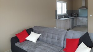 A modern living room with a grey sofa and red and white cushions, a kitchen with grey cabinets and a window, and a beige wall. - We all want to go to the Ardèche! It's great! (Gras)