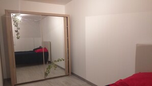 A bedroom with a wooden wardrobe, a bed with a black and red bedspread, and a small plant on the floor. - We all want to go to the Ardèche! It's great! (Gras)