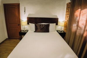 Quarto de hotel com porta de madeira, cama com roupa branca, duas luminárias de cabeceira e parede de tijolos decorativa.