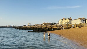 A coastal scene with a wooden pier extending into the sea, a sandy beach, and buildings in the background. - Oyster Stay House | By My Getaways (Whitstable)