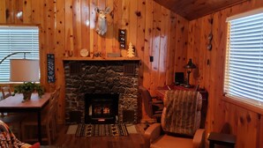 Interior acogedor de una cabaña con chimenea de piedra, paredes de madera, comedor y zona de estar cómoda.