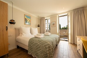 A hotel room with a bed, a desk, a balcony with a railing, and a painting on the wall.