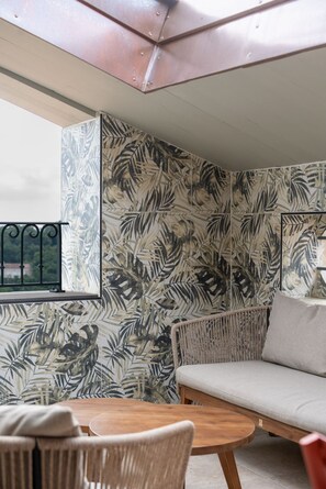 A room with a large window, a round wooden table, a sofa, and a wall with a tropical leaf pattern.