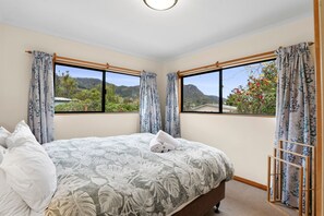 A bedroom with a bed, two windows with curtains, a view of greenery and mountains, and a ceiling light.
