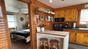 Interno di una baita in legno con cucina, zona pranzo con sgabelli, un letto e una ventola a soffitto.
