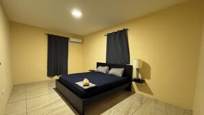 A bedroom with a bed, curtains, an air conditioning unit, a nightstand with a lamp, and tiled flooring. - ZEMMA Apartments Unit 2 (Willemstad)