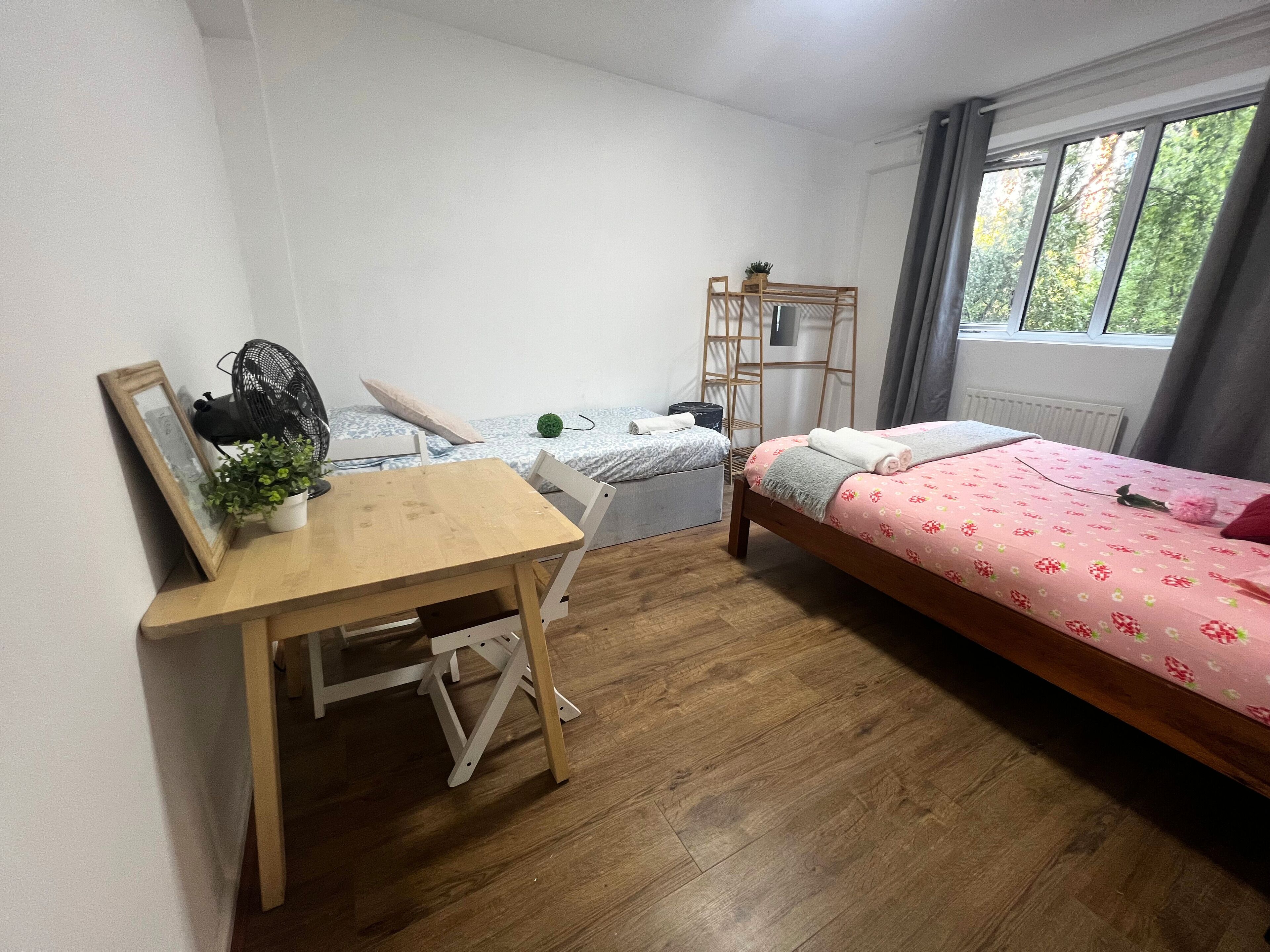 Een compacte, verzorgde slaapkamer met een houten bureau, een bed met een bloemenbedekking, een klein tafeltje met een plant en een raam met gordijnen.